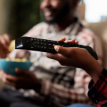 MUJER SOSTENIENDO CONTROL REMOTO DE TV CON HOMBRE AL FONDO